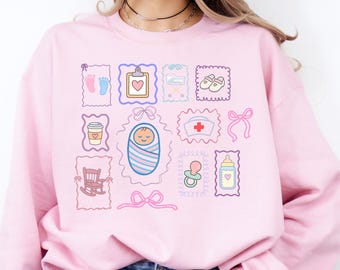 Könnte beinhalten: Hellrosa Sweatshirt mit einem Design zum Thema Baby. Das Design umfasst Babyfüße, ein Herz, ein in eine Decke gewickeltes Baby, einen Schnuller, eine Babyflasche und andere verwandte Bilder.