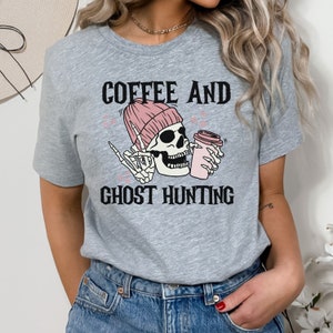 Op de afbeelding: Grijs T-shirt met een skelet dat een roze muts draagt en een roze koffiebeker vasthoudt. De tekst "Coffee and Ghost Hunting" staat op het T-shirt gedrukt.
