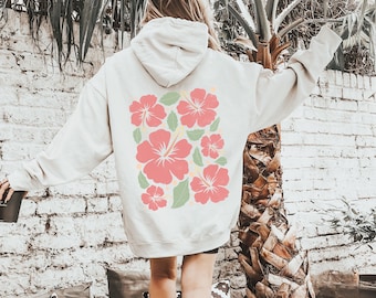May include: A white hoodie with a floral design on the back. The design features pink hibiscus flowers with green leaves and yellow centers.
