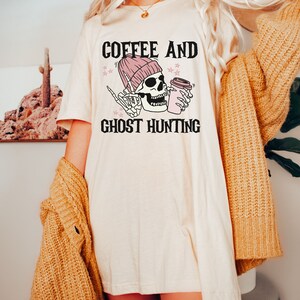 Op de afbeelding: Wit T-shirt met een zwarte en roze afbeelding van een skelet dat een roze muts draagt en een koffiebeker vasthoudt. De tekst "Coffee and Ghost Hunting" staat onder de afbeelding.