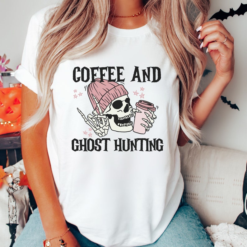 Op de afbeelding: Wit T-shirt met een roze en zwarte skeletgrafiek die een koffiebeker vasthoudt. De tekst "Coffee and Ghost Hunting" is onder de grafiek gedrukt.