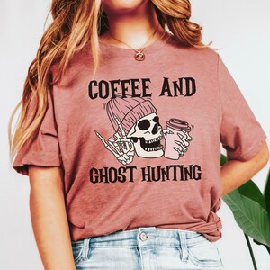 Op de afbeelding: Een roze T-shirt met de tekst "Coffee and Ghost Hunting" in zwart. Een cartoon-skelet met een roze muts houdt een roze koffiebeker vast.