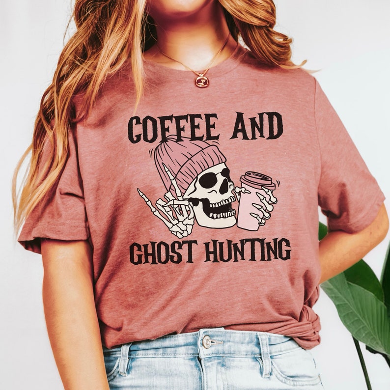 Op de afbeelding: Een roze T-shirt met de tekst "Coffee and Ghost Hunting" in zwart. Een cartoon-skelet met een roze muts houdt een roze koffiebeker vast.