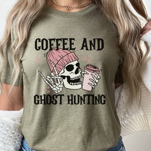 Op de afbeelding: Een groen T-shirt met de tekst "Coffee and Ghost Hunting" in zwart. Een cartoon-schedel met een roze muts houdt een roze koffiebeker vast.