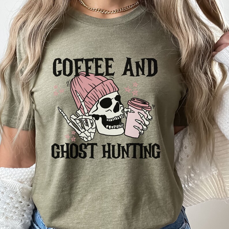 Op de afbeelding: Een groen T-shirt met de tekst "Coffee and Ghost Hunting" in zwart. Een cartoon-schedel met een roze muts houdt een roze koffiebeker vast.