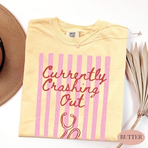 以下が含まれることがあります： バター色のTシャツ。赤色の筆記体で「Currently Crashing Out」と書かれ、ピンクの縦縞と聴診器のグラフィックがアクセントになっています。Comfort Colorsブランドのシャツです。
