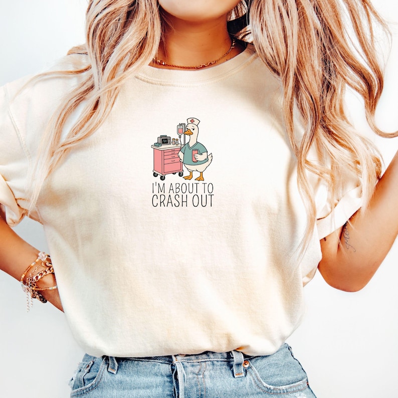 May include: Cream-colored t-shirt featuring a cartoon goose dressed as a nurse, holding a clipboard, and standing next to a pink medical cart. The text "I'M ABOUT TO CRASH OUT" is printed below the goose.