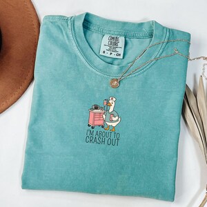 May include: Teal Comfort Colors t-shirt with a cartoon goose wearing a nurse's cap and scrubs, standing next to a pink medical cart. The text "I'M ABOUT TO CRASH OUT" is below the goose. A gold necklace is draped across the shirt.