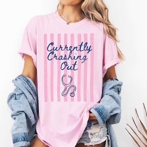 以下が含まれることがあります： ピンク色のTシャツ。ピンクのストライプと、紺色の「Currently Crashing Out」の文字がプリントされています。青い聴診器のグラフィックが下にあります。カジュアルなスタイルのシャツです。