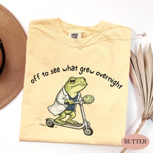 May include: Butter-colored t-shirt with a frog scientist graphic riding a scooter and the text "off to see what grew overnight." The frog wears a lab coat and holds a petri dish. A gold necklace with a star pendant is also visible.