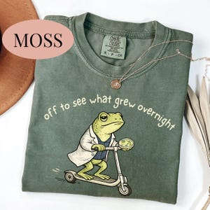 May include: Green t-shirt with the text "off to see what grew overnight" and a cartoon frog riding a scooter. The frog wears a lab coat and holds a petri dish. A silver necklace is draped across the shirt.