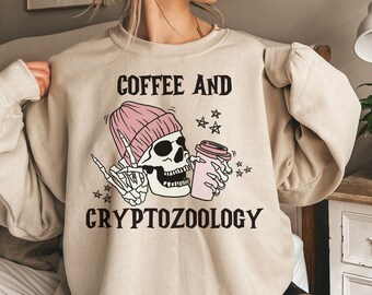 May include: A beige sweatshirt with a graphic print of a skull wearing a pink beanie and holding a coffee cup. The text "COFFEE AND CRYPTOZOOLOGY" is printed below the graphic.
