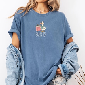 May include: A blue t-shirt with a graphic of a goose wearing a nurse's cap and scrubs, holding a coffee cup. A pink medical cart is next to the goose. The text "I'M ABOUT TO CRASH OUT" is below the graphic.
