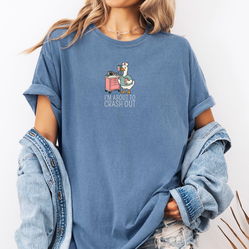 May include: A blue t-shirt with a graphic of a goose wearing a nurse's cap and scrubs, holding a coffee cup. A pink medical cart is next to the goose. The text "I'M ABOUT TO CRASH OUT" is below the graphic.