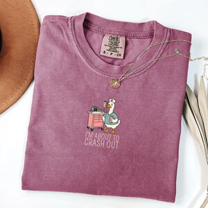 May include: A mauve Comfort Colors t-shirt featuring a cartoon goose in a nurse's uniform, standing next to a medical cart. The text "I'M ABOUT TO CRASH OUT" is below the graphic. A gold necklace and a brown hat are also visible.