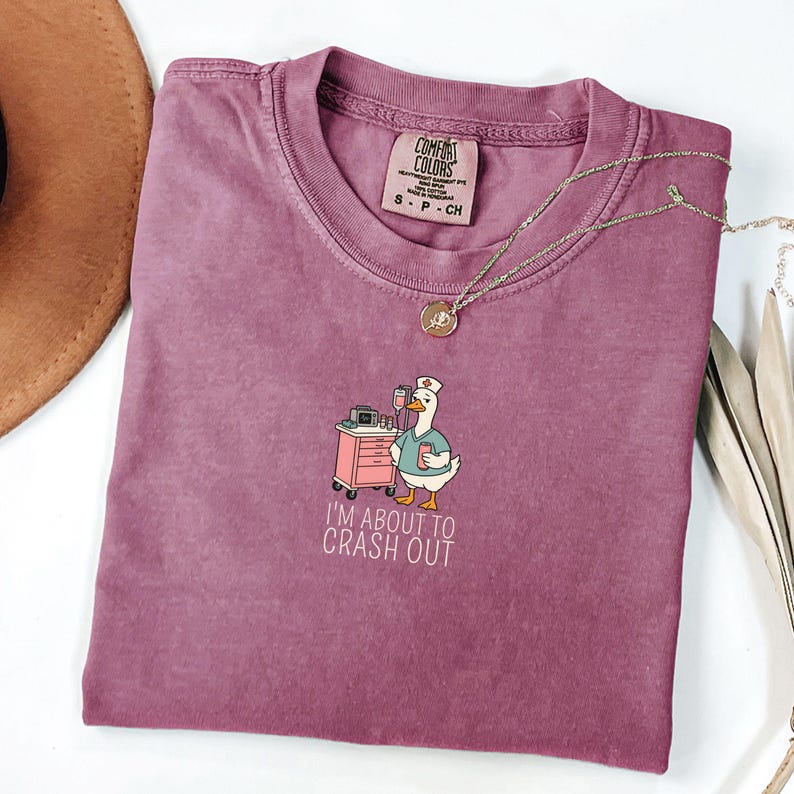 May include: A mauve Comfort Colors t-shirt featuring a cartoon goose in a nurse's uniform, standing next to a medical cart. The text "I'M ABOUT TO CRASH OUT" is below the graphic. A gold necklace and a brown hat are also visible.
