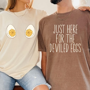 May include: Two t-shirts, one cream and one brown, with food-themed designs. The cream shirt has two egg illustrations, and the brown shirt has the text "JUST HERE FOR THE DEVILED EGGS" in white.