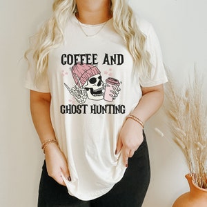 Op de afbeelding: Een wit T-shirt met een afbeelding van een skelet dat een roze muts draagt en een koffiebeker vasthoudt. De tekst "Coffee and Ghost Hunting" staat onder de afbeelding.