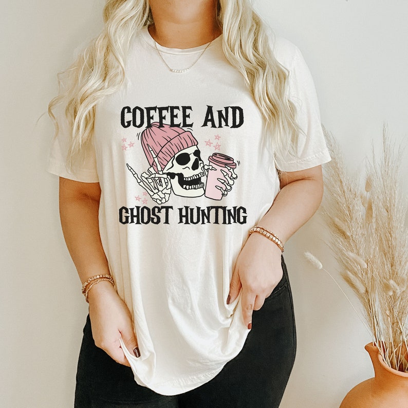 Op de afbeelding: Een wit T-shirt met een afbeelding van een skelet dat een roze muts draagt en een koffiebeker vasthoudt. De tekst "Coffee and Ghost Hunting" staat onder de afbeelding.