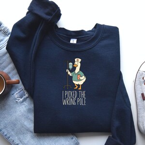 May include: Navy blue sweatshirt featuring a cartoon goose dressed as a nurse, holding an IV drip. The text "I PICKED THE WRONG POLE" is printed below the goose. The sweatshirt is made of a soft material.