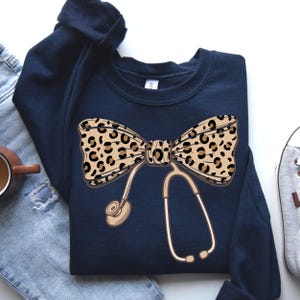 Könnte beinhalten: Marineblaues Sweatshirt mit Leopardenmuster-Schleife und Stethoskop-Design. Die Schleife ist beige mit schwarzen Punkten. Das Stethoskop ist beige mit einem goldfarbenen Ohrstück. Das Sweatshirt hat einen Rundhalsausschnitt.