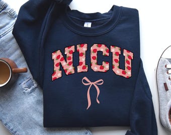 May include: A navy blue sweatshirt with a pink and white strawberry print. The text "NICU" is printed in white on the front of the sweatshirt. A pink bow is below the text.