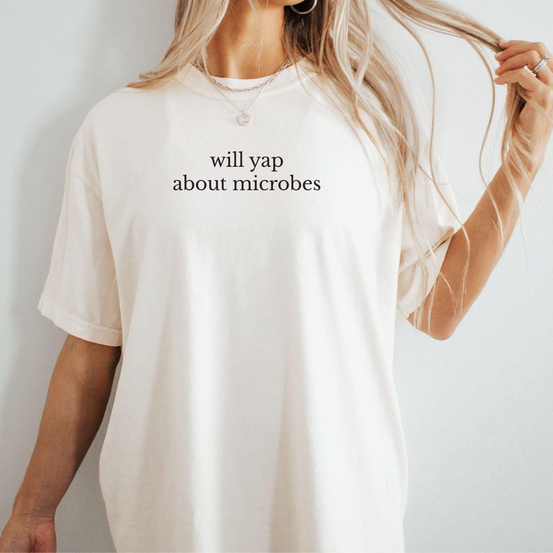 May include: Cream-colored t-shirt with the phrase "will yap about microbes" printed in black. The shirt has a relaxed fit and short sleeves. The text is centered on the front of the shirt.