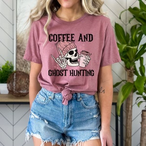 Op de afbeelding: Een roze T-shirt met de tekst "Coffee and Ghost Hunting" en een cartoon-skelet dat een roze muts draagt en een kopje koffie vasthoudt.