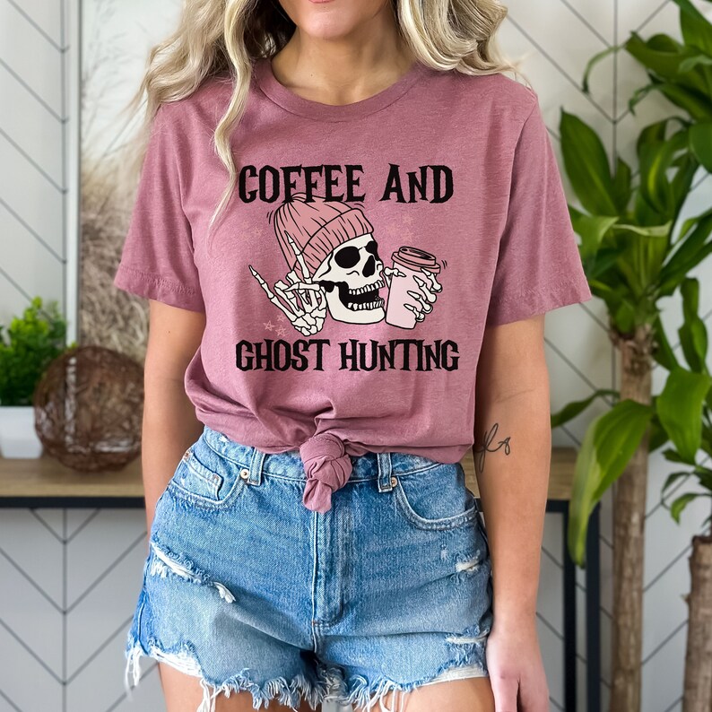 Op de afbeelding: Een roze T-shirt met de tekst "Coffee and Ghost Hunting" en een cartoon-skelet dat een roze muts draagt en een kopje koffie vasthoudt.