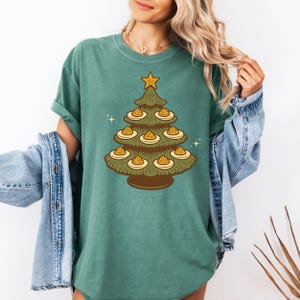 Peut inclure: T-shirt vert avec un motif d'arbre de Noël de style rétro. L'arbre marron est orné de décorations jaunes et marron, surmonté d'une étoile dorée. Le t-shirt a des manches courtes et un col rond.