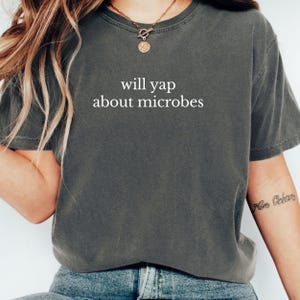 May include: Dark grey t-shirt with the phrase "will yap about microbes" printed in white. The shirt has short sleeves and a crew neck. The shirt is made of a soft, comfortable material.