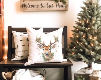 May include: A decorative white pillow with a watercolor illustration of a deer with antlers adorned with greenery and red berries. A second pillow with an arrow pattern is visible. A wooden sign above reads "Welcome to Our Home". A small Christmas tree is also in the image.