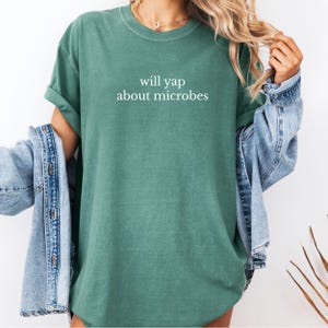 May include: A teal-colored t-shirt with the phrase "will yap about microbes" printed in white. The shirt has a crew neck and short sleeves. A denim jacket is draped over the shoulders.