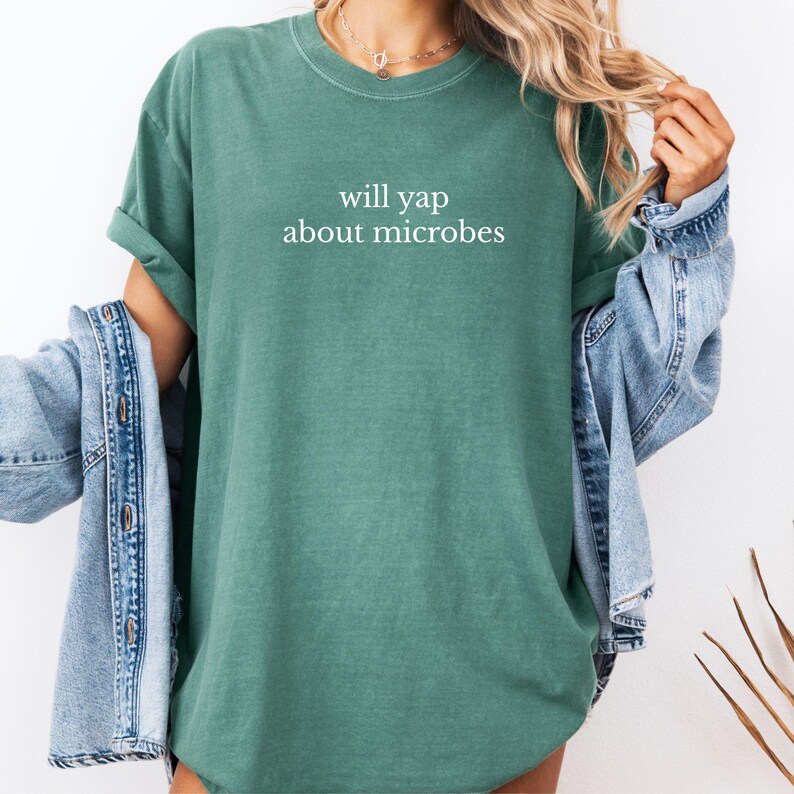 May include: A teal-colored t-shirt with the phrase "will yap about microbes" printed in white. The shirt has a crew neck and short sleeves. A denim jacket is draped over the shoulders.