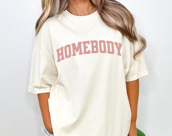 Op de afbeelding: Een oversized t-shirt in crèmekleur met het woord "HOMEBODY" gedrukt in een roze boog.