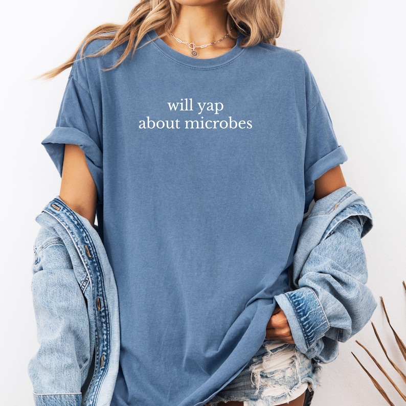 May include: A blue t-shirt with the text "will yap about microbes" in white. The shirt has rolled-up sleeves and is paired with a denim jacket. The overall style is casual and relaxed.