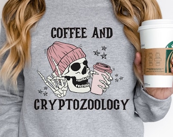 May include: Gray sweatshirt with a skeleton wearing a pink beanie holding a coffee cup. The text on the shirt reads "Coffee and Cryptozoology" with stars around the skeleton.