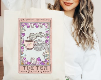 Puede incluir: Una bolsa de tela blanca con un diseño de carta de tarot rosa y gris. La carta presenta una mano esquelética sosteniendo una taza de té humeante rodeada de cristales. El texto "THE TEA" está impreso en la carta.