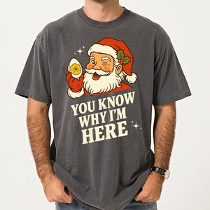 Puede incluir: Camiseta gris oscuro con una ilustración retro de Papá Noel guiñando un ojo y sosteniendo un huevo relleno. El texto "YOU KNOW WHY I'M HERE" está impreso debajo, con acentos de estrellas. La camiseta tiene un cuello redondo clásico.