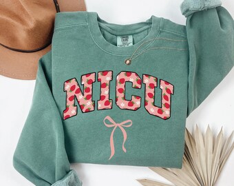 May include: A green sweatshirt with a pink and white strawberry print that says "NICU" in a collegiate font with a pink bow underneath.