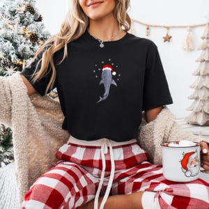 May include: A black t-shirt with a white shark wearing a red Santa hat. The shark is surrounded by white snowflakes. The person wearing the shirt is also wearing red and white plaid pajama pants.
