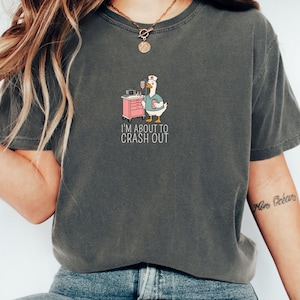 May include: Dark gray t-shirt featuring a cartoon goose in a nurse's uniform, standing next to a pink medical cart. The goose holds a clipboard and a drink. The text "I'M ABOUT TO CRASH OUT" is below the goose.