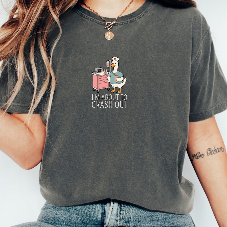 May include: Dark gray t-shirt featuring a cartoon goose in a nurse's uniform, standing next to a pink medical cart. The goose holds a clipboard and a drink. The text "I'M ABOUT TO CRASH OUT" is below the goose.