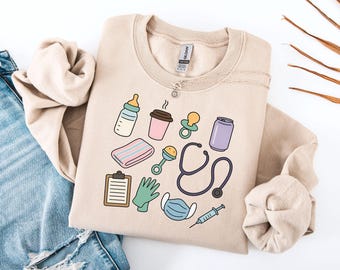 Könnte beinhalten: Beiges Sweatshirt mit farbenfrohen Illustrationen von Baby- und medizinischen Artikeln. Das Design umfasst eine Babyflasche, eine Kaffeetasse, einen Schnuller, eine Rassel, ein Stethoskop, eine medizinische Maske und eine Spritze. Das Sweatshirt wird mit einer blauen Jeans kombiniert.