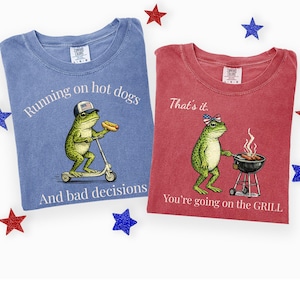 Könnte beinhalten: Zwei Comfort Colors T-Shirts. Eines ist blau mit einem Frosch auf einem Roller und dem Text "Running on hot dogs and bad decisions." Das andere ist rot mit einem Frosch am Grill und dem Text "That's it. You're going on the GRILL."