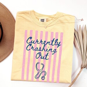 以下が含まれることがあります： 明るい黄色のTシャツ。テキスト「Currently Crashing Out」は濃い青色の筆記体で、ピンクの縦縞模様の上に配置されています。青い聴診器のグラフィックが下にあります。このシャツはComfort Colorsブランドです。