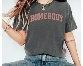 Op de afbeelding: Een donkergrijs T-shirt met het woord "HOMEBODY" in roze op de voorkant. Het shirt wordt gedragen door een persoon met lang blond haar en een gouden ketting.