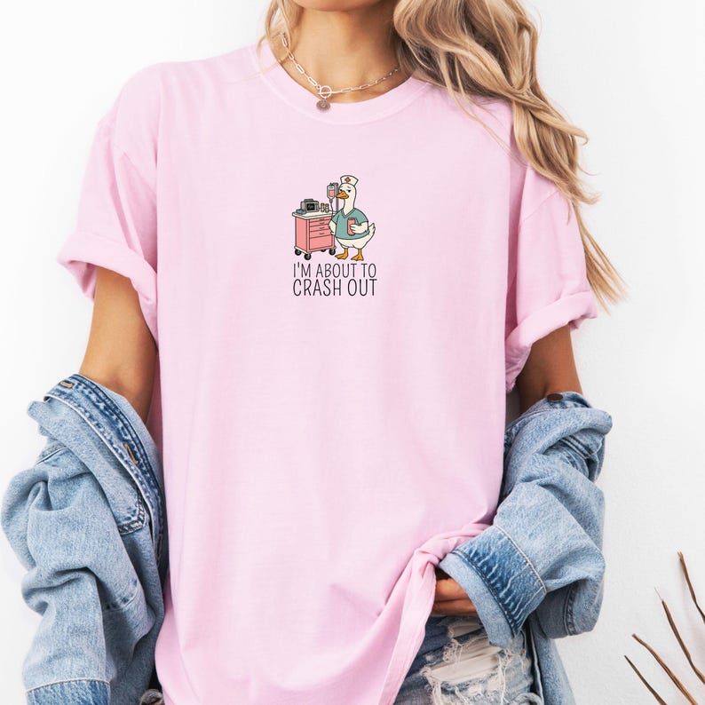 May include: A light pink t-shirt featuring a cartoon goose in a nurse's hat, standing next to a medical cart. The goose is holding a clipboard and a cup. The text "I'M ABOUT TO CRASH OUT" is printed below the goose.