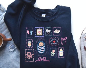 Könnte beinhalten: Marineblaues Sweatshirt mit Baby-Illustrationen in Pastellfarben. Das Design umfasst Babyfüße, ein Herz, ein Baby in einer Windel, eine Flasche und einen Schnuller. Das Sweatshirt ist ein bequemer, lässiger Stil.