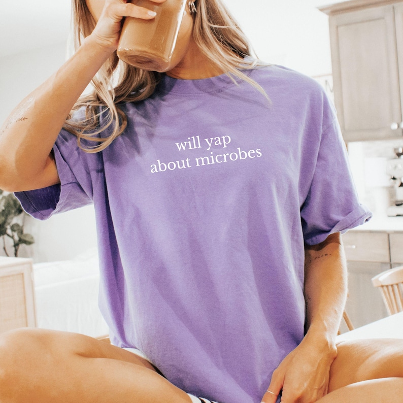 May include: A lilac-colored t-shirt with the text "will yap about microbes" in white. The shirt has short sleeves and a relaxed fit. The person in the image is drinking from a glass.
